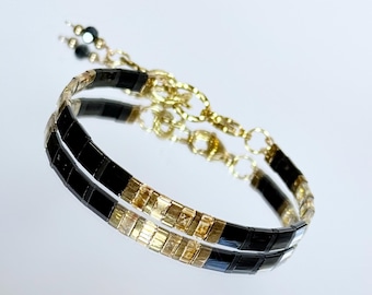 May include: A delicate bracelet with alternating black and gold rectangular beads. The bracelet is connected by a gold chain and features small black beads at the end. The bracelet is displayed on a reflective surface.
