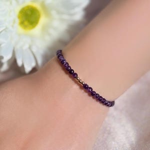 May include: A delicate bracelet made of small, round, deep purple amethyst beads, interspersed with gold-coloured beads. The bracelet is worn on a wrist. A white flower is visible in the background.