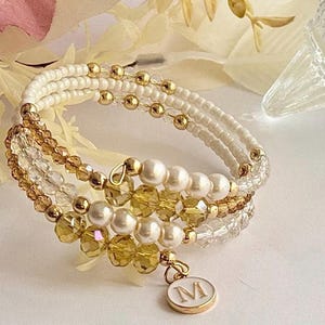 May include: A multi-strand wrap bracelet featuring white, gold, and clear beads. The bracelet includes gold-colored accents, faux pearls, and a charm with the letter "M". The bracelet is designed to wrap around the wrist multiple times.