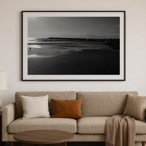 May include: A black and white photograph of a beach with a long, low wall extending into the water. The sand is wet and there are ripples in the water.