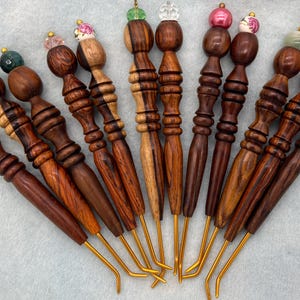 May include: A collection of eleven handcrafted crochet hooks featuring turned wooden handles in various shades of brown, ranging from light to dark wood grain. Each handle is topped with a decorative bead in colors like pink, blue, green, and clear.