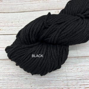 May include: A large skein of black yarn, tightly wound and textured, rests on a light wood surface. The word "BLACK" is printed in white on the yarn. This yarn is suitable for various knitting and crochet projects.