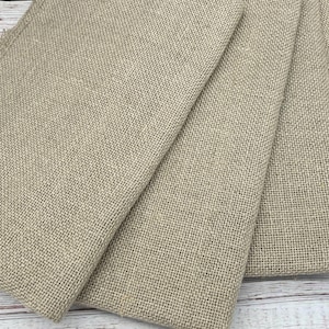 May include: Close-up of several folded burlap fabric pieces. The fabric is a natural tan color with a coarse, woven texture. The edges of the fabric are frayed, and the pieces are arranged on a light-colored wooden surface.