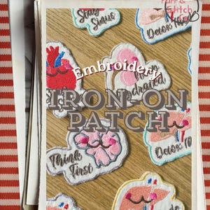 May include: A collection of embroidered iron-on patches featuring anatomical designs including a heart, brain, and liver with smiling faces and text such as Think First and Stay Sinus. The patches are white with colorful thread details.