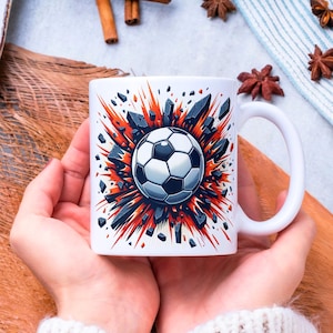 May include: White ceramic mug featuring a soccer ball design. The soccer ball is in the center of an explosion of red and gray shards. The mug has a white handle and is held by a person.