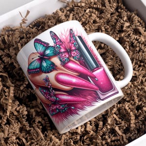 Könnte beinhalten: Weiße Keramik-Tasse mit einem Nageldesign. Das Design umfasst lange, pinkfarbene Nägel, Schmetterlinge mit türkisfarbenen und rosa Flügeln und eine Flasche pinkfarbenen Nagellacks. Die Tasse befindet sich in einer Geschenkbox mit braunem Verpackungsmaterial.