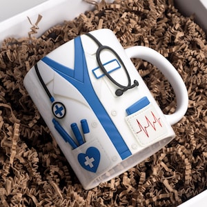 May include: White ceramic mug decorated to resemble a doctor's coat, complete with a stethoscope, blue pens, and a heart-shaped pocket with a medical cross. The mug also features a red EKG line and is presented in a gift box with brown paper filler.