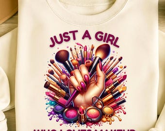 Puede incluir: Sudadera crema con un gráfico vibrante de una mano sosteniendo brochas de maquillaje y pintalabios. El texto "JUST A GIRL WHO LOVES MAKEUP" está en morado. El diseño incluye varios productos de maquillaje.