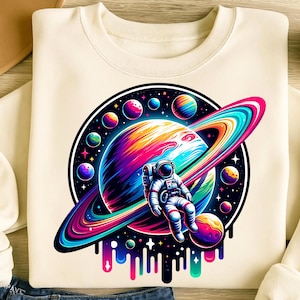 May include: Cream-colored sweatshirt featuring a vibrant graphic of a colorful planet with rings, surrounded by smaller planets. An astronaut floats in front of the planet. The design includes a black border and colorful drips at the bottom.