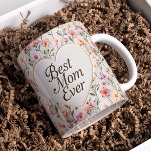 Op de afbeelding: Witte keramische mok met een bloemenpatroon en de tekst "Best Mom Ever" in een hartvorm. De mok heeft roze, perzik en gele bloemen. De mok zit in een witte doos met bruine papieren vulling.