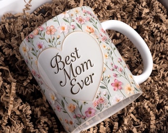 May include: White ceramic mug with a floral design and the text "Best Mom Ever" inside a heart shape. The mug features pink, peach, and yellow flowers. The mug is inside a white box with brown paper filler.
