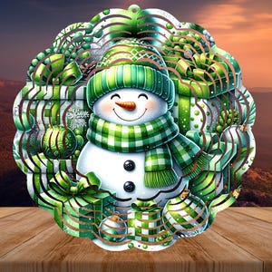 May include: A whimsical Christmas decoration featuring a snowman wearing a green and white plaid scarf and hat. The snowman is surrounded by green gift boxes and ornaments. The design is set against a blurred mountain backdrop.