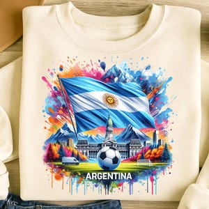 Op de afbeelding: Crèmekleurige sweatshirt met een levendige afbeelding van de Argentijnse vlag, een voetbal en een stadsgezicht. Het ontwerp bevat kleurrijke verfspatten en het woord "ARGENTINA". De sweatshirt is gemaakt van zacht materiaal.