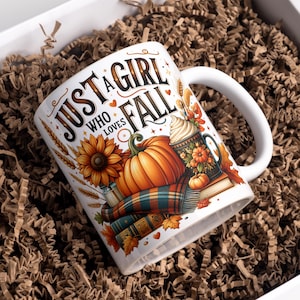 Op de afbeelding: Witte keramische mok met de tekst "JUST A GIRL WHO LOVES FALL" en herfstillustraties. Het ontwerp bevat een zonnebloem, pompoenen, een geruite sjaal en een decoratieve mok met slagroom. De mok zit in een geschenkverpakking.