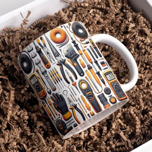 May include: White ceramic mug featuring a detailed illustration of various tools, including pliers, screwdrivers, and multimeters. The tools are in black, orange, and gray, set against a white background. The mug has a white handle and is presented in a box with brown packing material.