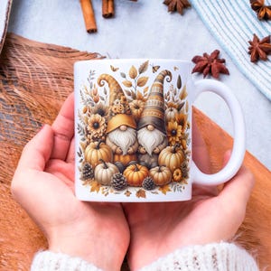 May include: White ceramic mug featuring a fall-themed design with two gnomes, pumpkins, and autumn foliage in shades of orange, brown, and yellow. The mug has a white handle and is held by a person.