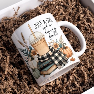 May include: A white ceramic mug featuring a graphic of an iced coffee in a plastic cup, a black and white plaid scarf, stacked books, pumpkins, wheat stalks, and autumn leaves. The text "JUST A GIRL who loves fall" is printed in black script.
