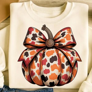 May include: Cream-colored sweatshirt featuring a pumpkin design. The pumpkin is adorned with a large bow, showcasing a pattern of orange, red, and brown spots. The pumpkin has a brown stem and a decorative center. The design is centered on the front of the sweatshirt.