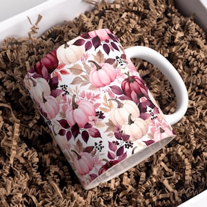 May include: A white ceramic mug featuring a repeating pattern of light pink, cream, and deep burgundy pumpkins interspersed with dark purple leaves, small berries, and delicate floral sprigs. The mug has a simple white handle and a smooth finish.