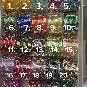 K&ouml;nnte beinhalten: Eine Sammlung farbenfroher Glasarmreifen in einem transparenten Kunststoffbeh&auml;lter. Die Armreifen sind in verschiedenen Farben erh&auml;ltlich, darunter Gelb, Gr&uuml;n, Blau, Rot und Lila. Jeder Armreif ist mit kleinen, goldfarbenen Verzierungen versehen. Die Armreifen sind von 1 bis 24 nummeriert.
