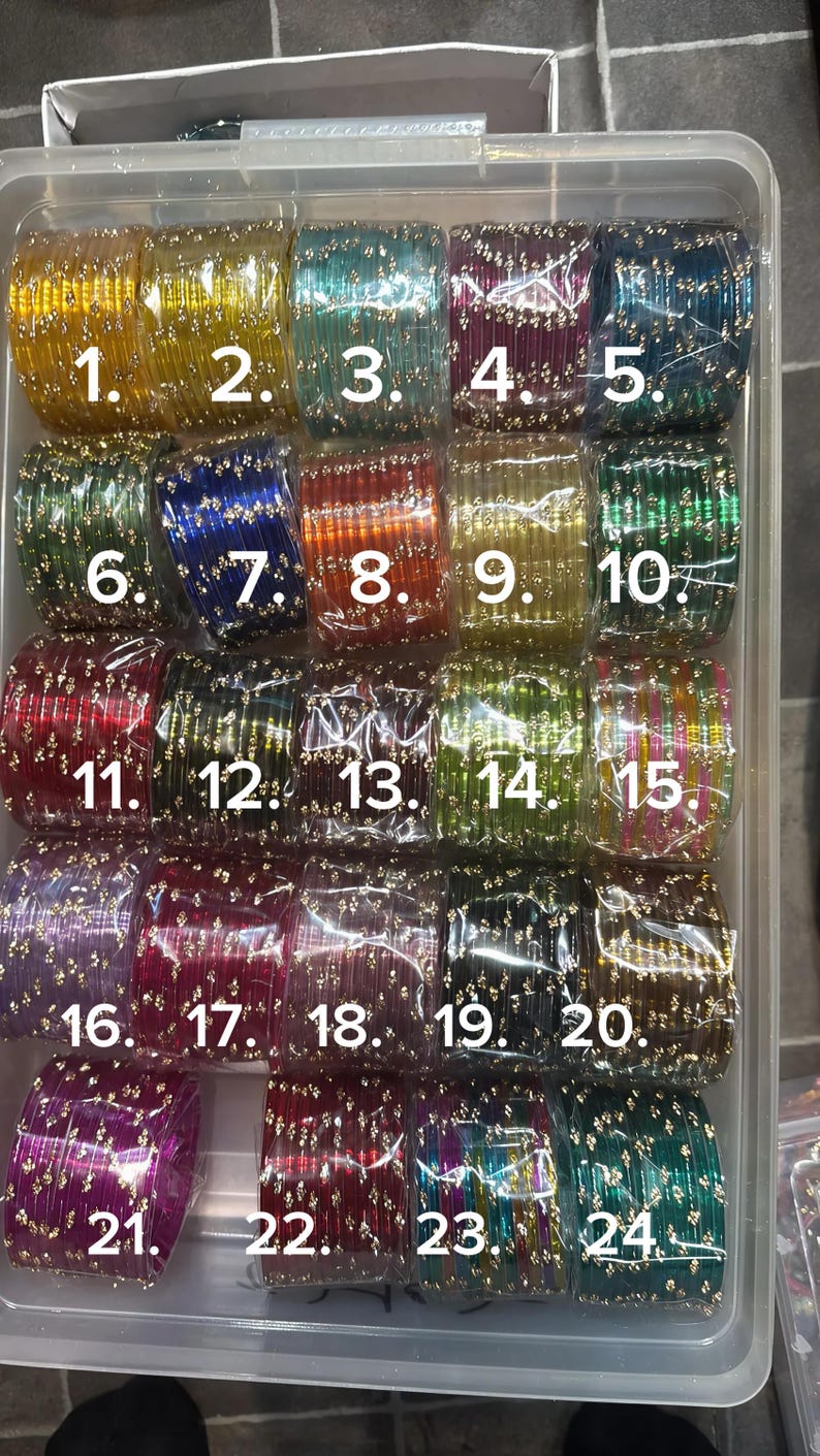 K&ouml;nnte beinhalten: Eine Sammlung farbenfroher Glasarmreifen in einem transparenten Kunststoffbeh&auml;lter. Die Armreifen sind in verschiedenen Farben erh&auml;ltlich, darunter Gelb, Gr&uuml;n, Blau, Rot und Lila. Jeder Armreif ist mit kleinen, goldfarbenen Verzierungen versehen. Die Armreifen sind von 1 bis 24 nummeriert.