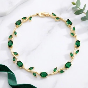 May include: A gold-toned bracelet featuring a vine-like structure adorned with multiple oval-shaped, deep green gemstones. The design incorporates small, leaf-shaped green accents along the metal band, secured with a rectangular clasp closure.