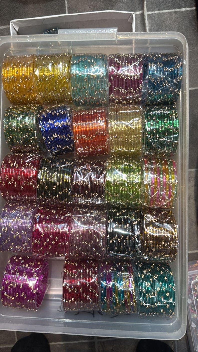 K&ouml;nnte beinhalten: Ein durchsichtiger Kunststoffbeh&auml;lter gef&uuml;llt mit mehreren Stapeln bunter Armreifen. Die Armreifen sind in verschiedenen Farben erh&auml;ltlich, darunter Gelb, Gr&uuml;n, Blau, Rot und Lila, und sind mit kleinen, goldfarbenen Verzierungen versehen.