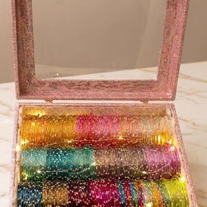 May include: A pink glitter-covered box with a clear lid, filled with rows of colorful glass bangles. The raindrops bangles are in shades of yellow, green, blue, pink, and red. Tiny fairy lights are strung throughout the bangles.