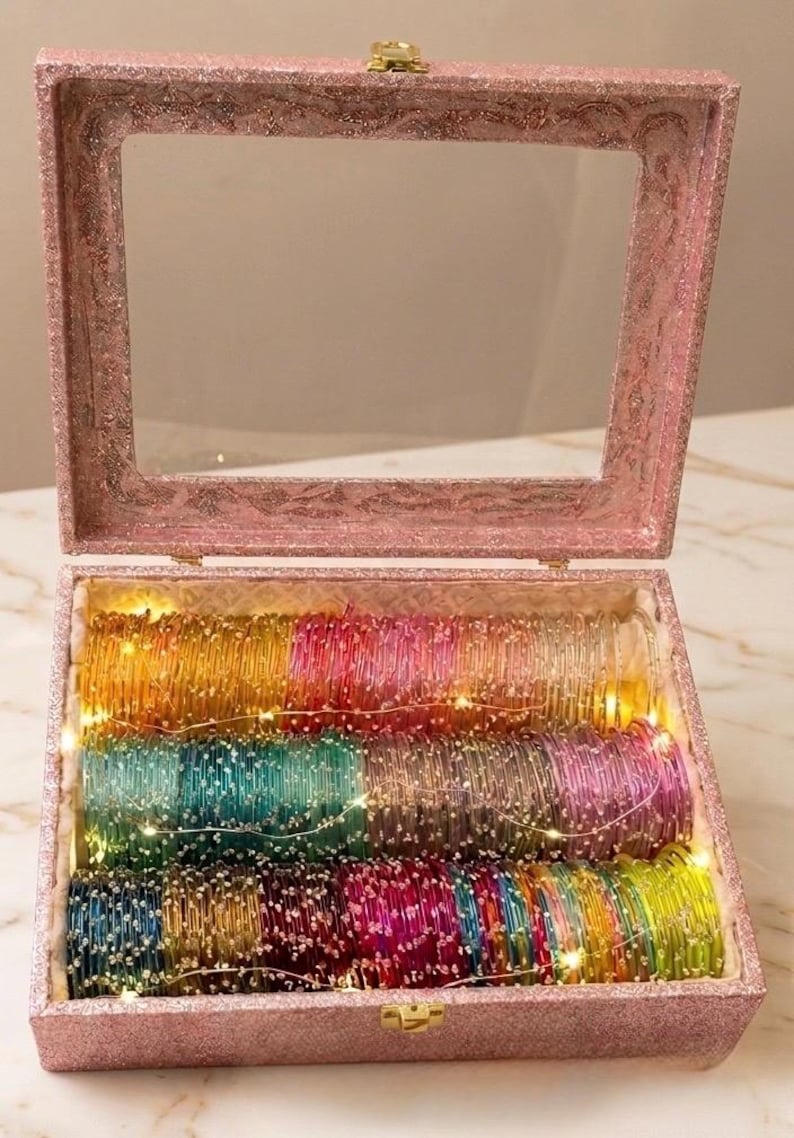 May include: A pink glitter-covered box with a clear lid, filled with rows of colorful glass bangles. The raindrops bangles are in shades of yellow, green, blue, pink, and red. Tiny fairy lights are strung throughout the bangles.