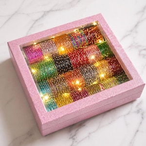 May include: A pink glitter box containing a collection of colourful bangles. The bangles are arranged in rows and lit by small, warm white lights. The bangles are in shades of pink, green, gold, and red.