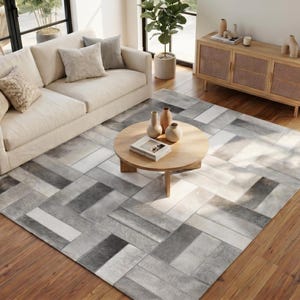 May include: A rectangular area rug featuring a geometric patchwork pattern of rectangular tiles in varying shades of gray, white, and charcoal. The rug is laid out on a wooden floor, partially covered by a round wooden coffee table.