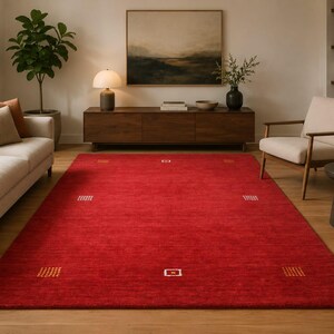 Puede incluir: Una alfombra roja rectangular grande con un patrón geométrico en un salón. La alfombra presenta pequeños diseños cuadrados y rectangulares en blanco y dorado. La alfombra está colocada sobre un suelo de madera, con un sofá y una silla cerca. Mide unos 300 cm x 200 cm.