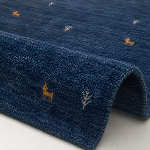 May include: A deep blue area rug with a textured pile surface featuring small, embroidered yellow deer and white tree motifs scattered across the fabric. The rug is folded over at one corner, revealing the thick, tufted construction and finished edge.
