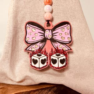 Könnte beinhalten: Ein rosa schleifenförmiger Anhänger mit goldenen Herzakzenten und zwei Würfeln mit Herzsymbolen. Der Anhänger ist an einer Kette aus rosa und orangefarbenen Perlen befestigt. Die Würfel sind von rotem Glitzer umgeben.