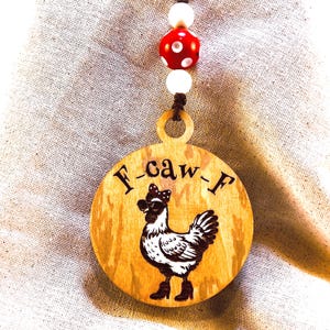 May include: A wooden pendant with a rooster illustration and the text "F-caw-F". The pendant is round with a brown cord and red and white beads. The rooster is wearing a bow and boots.