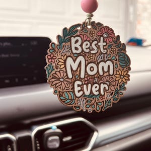 Puede incluir: Un adorno de coche de madera con las palabras "Best Mom Ever" en una fuente decorativa. El adorno está rodeado de diseños florales y de hojas en tonos rosa, verde y marrón. Una cuenta rosa cuelga de la parte superior.