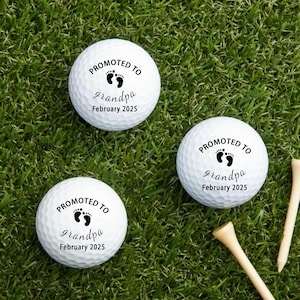 May include: Three white golf balls with the text "PROMOTED TO grandpa February 2025" in black. The golf balls are on green grass with two wooden golf tees.