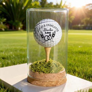 Op de afbeelding: Een golfbal met de namen "Lucas & Charlotte" en de datum "8.2026 December" erop gedrukt, tentoongesteld in een glazen cilinder. De bal rust op een houten tee boven groen mos, met een kurken basis.