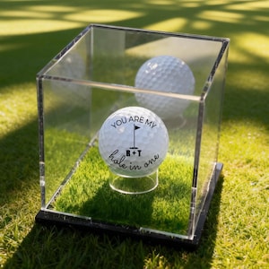 Pode incluir: Uma bola de golfe com o texto "You are my hole in one" dentro de uma caixa de exibição de acrílico transparente. A bola de golfe está em um pequeno suporte transparente, rodeada por grama verde artificial. Outra bola de golfe é visível no fundo.
