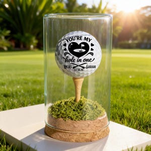 Könnte beinhalten: Ein Golfball mit dem Text "YOU'RE MY hole in one" in einer Glasvitrine. Der Golfball befindet sich auf einem Tee, umgeben von grünem Moos. Die Namen "LUCAS" und "SARAH" stehen unter dem Text, zusammen mit dem Datum "02.14.26".