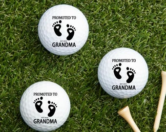 Puede incluir: Tres pelotas de golf blancas con el texto "PROMOTED TO GRANDMA" y huellas de bebé en negro. También se incluye el texto "EST 2025". Las pelotas de golf están sobre una superficie de hierba verde con tees de golf de madera.