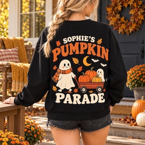 Op de afbeelding: Een zwart sweatshirt met lange mouwen en een opdruk op de achterkant met de tekst 'SOPHIE'S PUMPKIN PARADE'. Het ontwerp toont een geest met een geruite sjaal die een rode wagen vol pompoenen, herfstbladeren en een maansikkel voorttrekt.