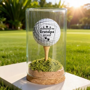 May include: A golf ball with the text "Promoted to Grandpa EST. 2025" inside a clear glass cylinder. The golf ball sits on a wooden tee, atop a bed of green moss, and a cork base. The background is a green lawn.