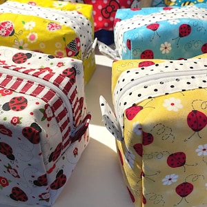 May include: Several fabric cosmetic bags with ladybug and floral patterns. The bags are in various colors, including yellow, blue, and white, and have white zippers and small fabric loops. The bags are likely handmade and suitable for storing makeup or toiletries.