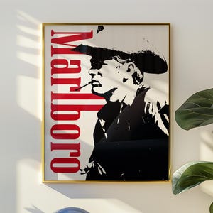 Op de afbeelding: Ingelijste poster met een zwart-wit silhouet van een rokende cowboy, met het woord "Marlboro" in rode verticale tekst. De poster zit in een gouden lijst, gemonteerd op een witte muur. Een blauwe lamp en een plant staan op de voorgrond.