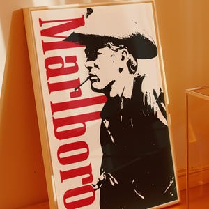 Op de afbeelding: Een ingelijste poster met een zwart-wit silhouet van een persoon met een cowboyhoed en een sigaret. Het woord "Marlboro" staat verticaal in het rood gedrukt. De poster zit in een lichtgekleurde lijst.
