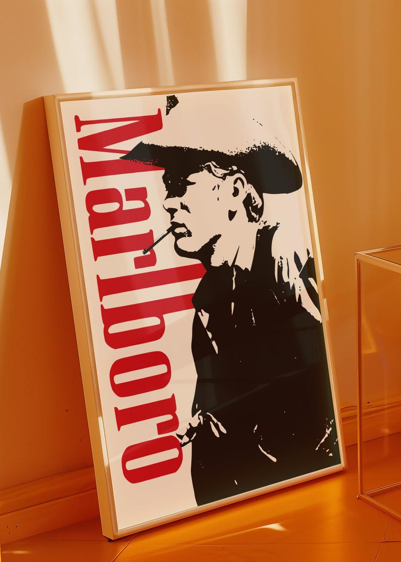 Op de afbeelding: Een ingelijste poster met een zwart-wit silhouet van een persoon met een cowboyhoed en een sigaret. Het woord "Marlboro" staat verticaal in het rood gedrukt. De poster zit in een lichtgekleurde lijst.