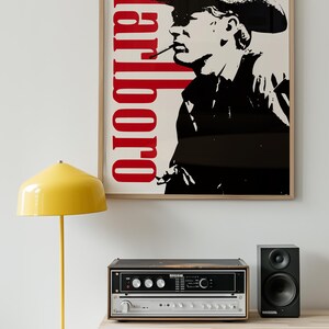 Op de afbeelding: Een ingelijste poster met het woord "Marlboro" in het rood en een zwart-wit afbeelding van een rokende cowboy. Een gele lamp en stereo-apparatuur staan op een gele kast. Rechts staat een zwarte luidspreker.