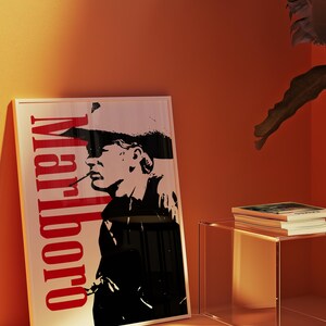 Op de afbeelding: Een ingelijste poster met een zwart-wit silhouet van een rokende persoon, met het woord "Marlboro" in het rood verticaal weergegeven. De poster leunt tegen een oranje muur, met een doorzichtige bijzettafel en een stapel boeken.