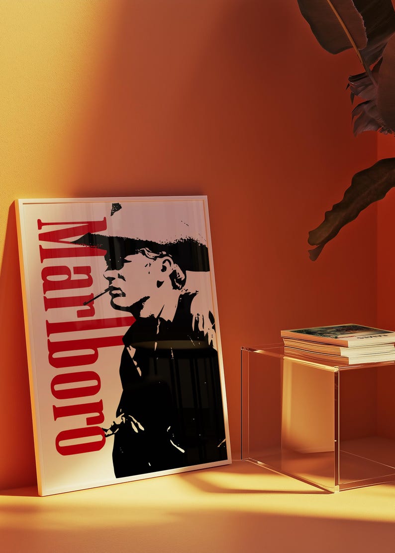 Op de afbeelding: Een ingelijste poster met een zwart-wit silhouet van een rokende persoon, met het woord "Marlboro" in het rood verticaal weergegeven. De poster leunt tegen een oranje muur, met een doorzichtige bijzettafel en een stapel boeken.